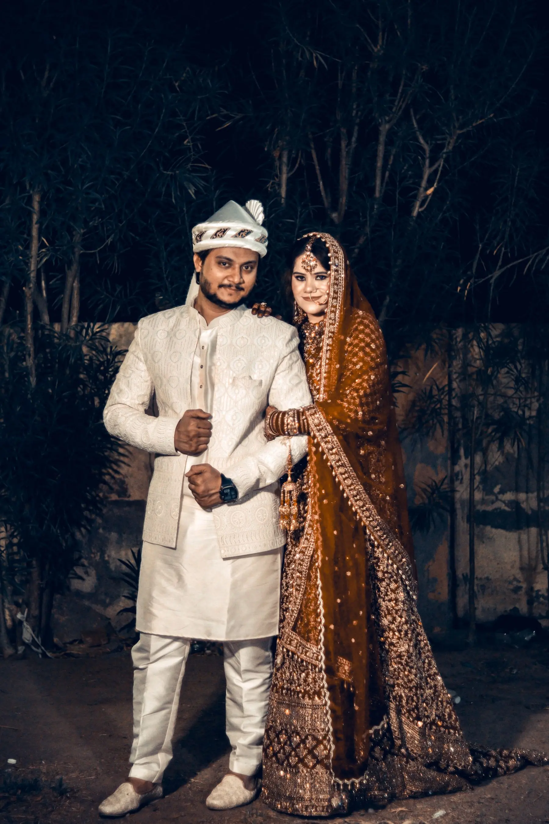 Indian bride and groom in traditional wedding attire photographed together outdoors.
